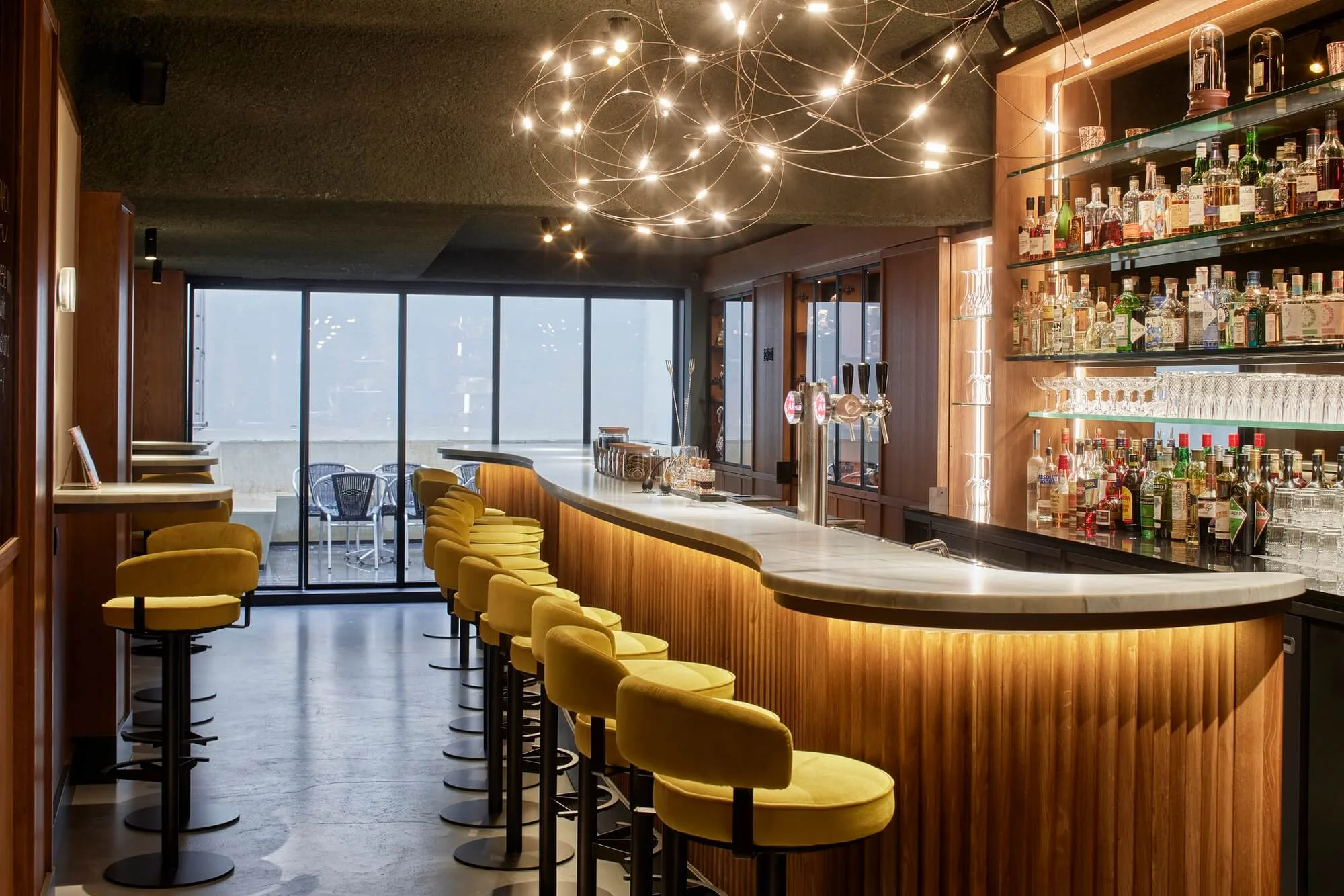 Breed zicht op de bar vanaf de andere kant, met warme vloerreflecties en de mosterdgele krukken